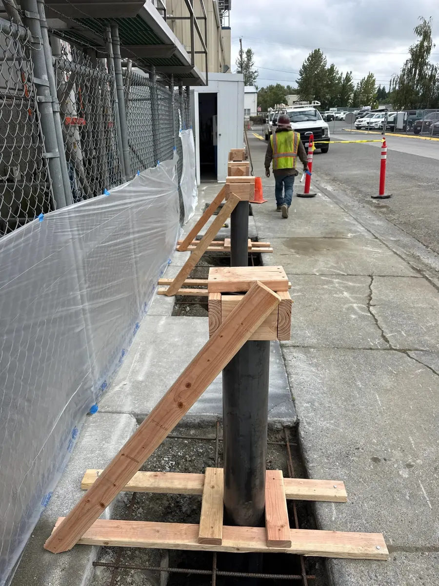 Row of steel pipe bollards supported with wood bracing along a concrete lane beside an industrial facility fence, crew member walking in background