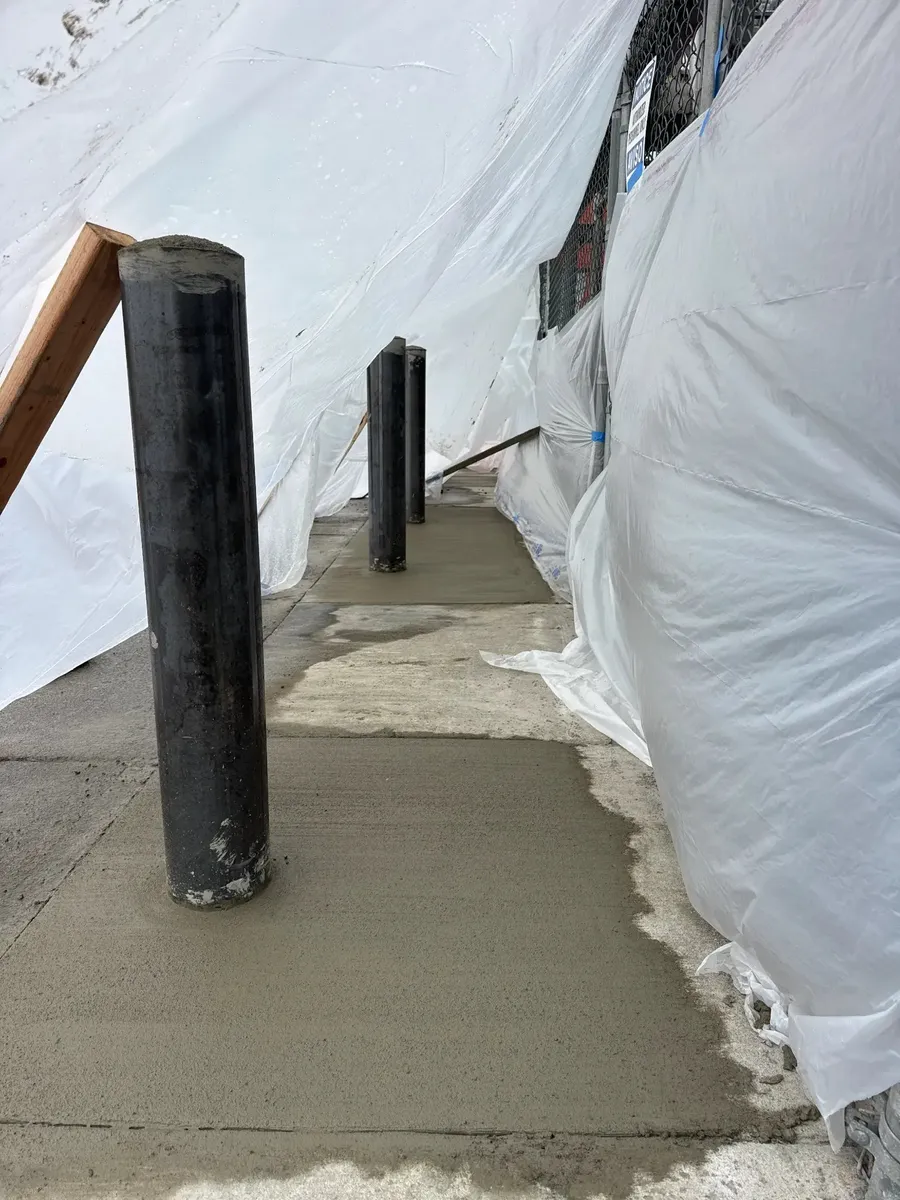 Steel bollards with fresh concrete placed around footings and plastic sheeting protecting the adjacent chain-link fence during curing