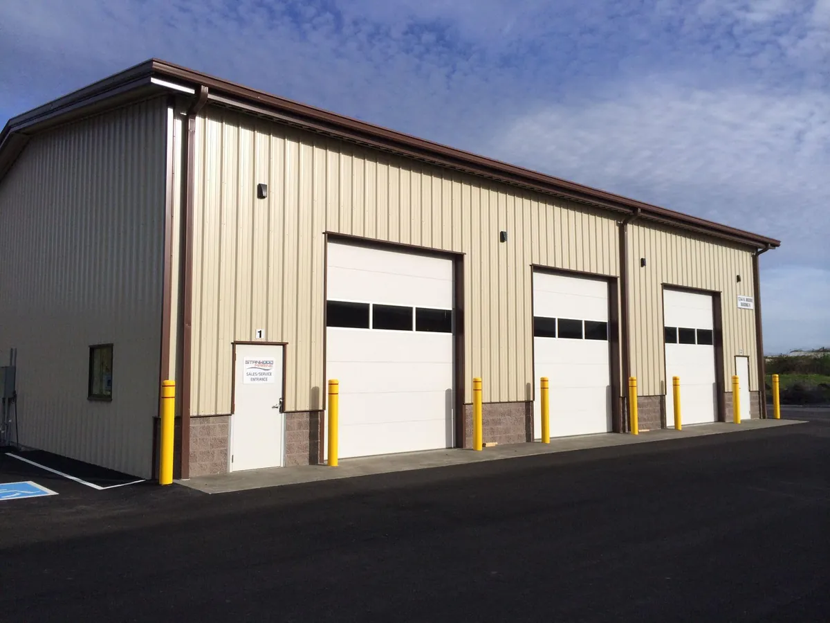 Completed metal commercial building with three overhead doors, paved vehicle access, and yellow protective bollards
