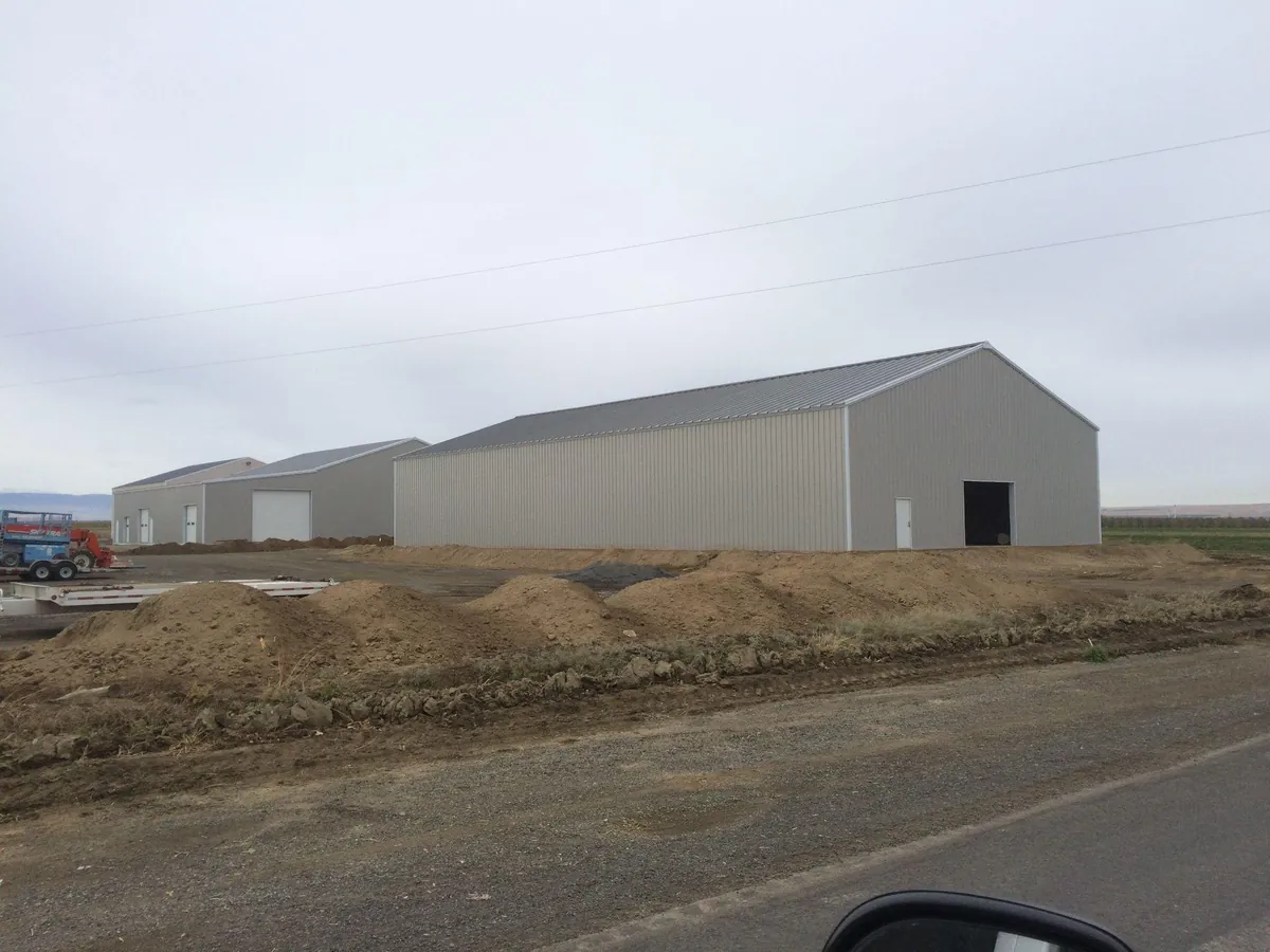 Completed light-gray steel buildings on an agricultural or light-industrial property with site grading in the foreground