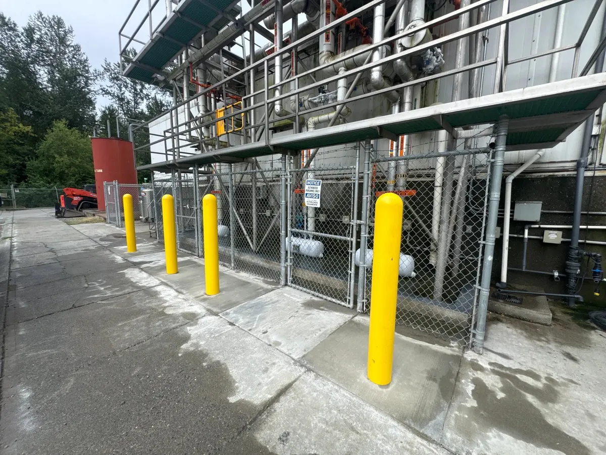 Completed safety-yellow steel bollards installed along a concrete lane protecting the chain-link fence and mechanical equipment at an industrial facility