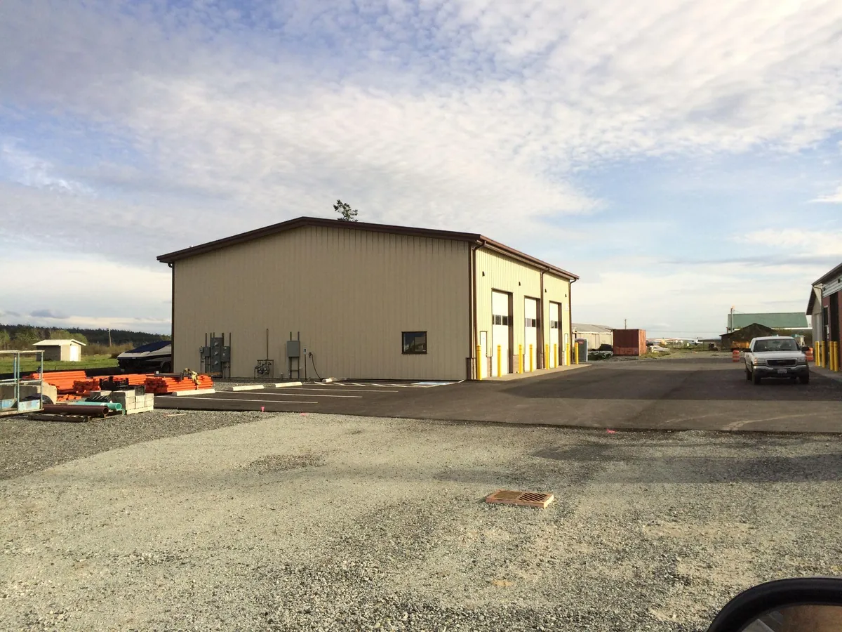 Completed metal commercial building with overhead doors, paved parking, and vehicle access beside a gravel work area