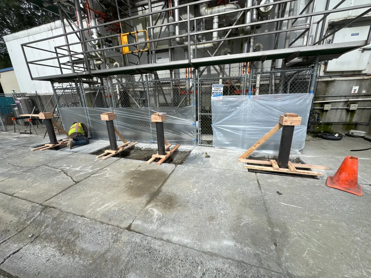 Multiple steel bollards set in footings with wood bracing along an industrial facility's chain-link fence with protective plastic sheeting over equipment