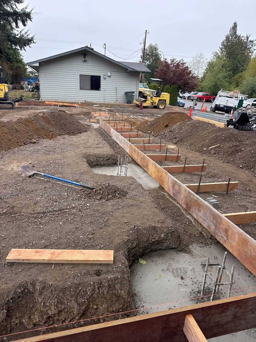 Excavated foundation runs, timber forms, and exposed reinforcement show the project moving through the foundation-preparation stage.
