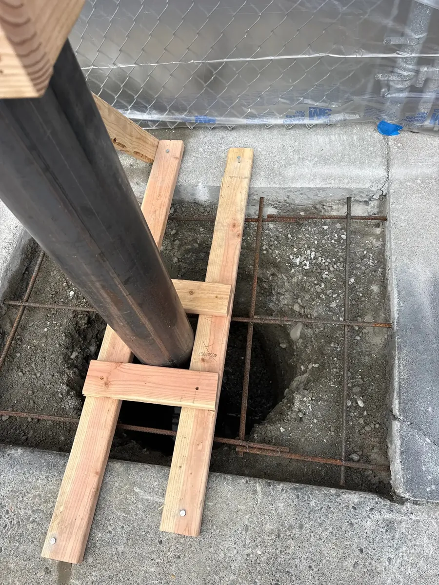 Close-up detail of a steel pipe bollard set in an excavated concrete footing with rebar reinforcement grid and dimensional lumber H-frame bracing holding it plumb