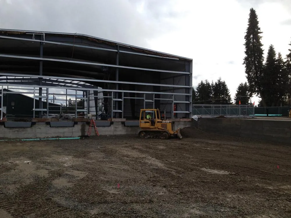 Steel building construction site with a tracked dozer grading soil beside open framed access bays and concrete foundation walls