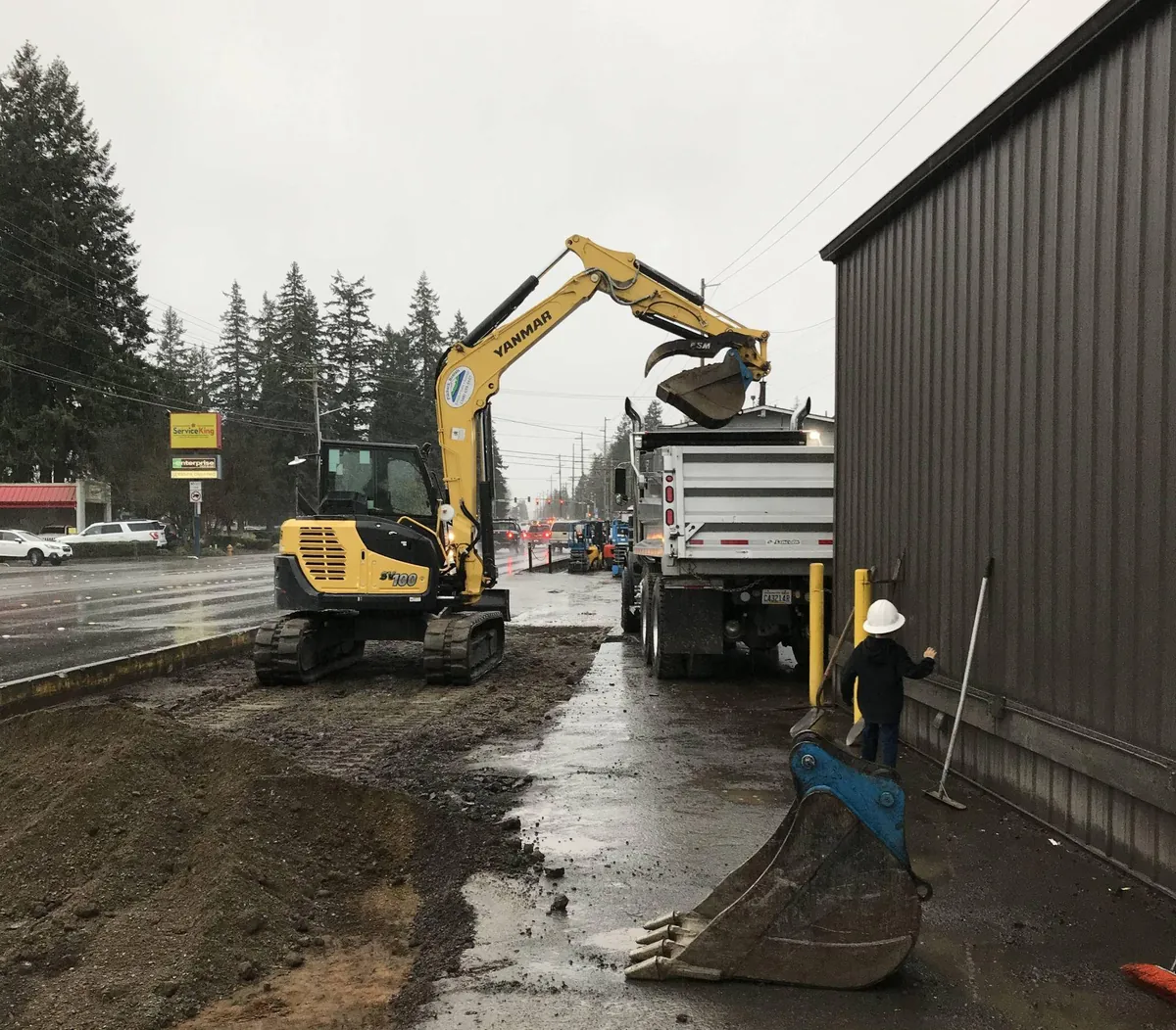 Excavator performing wet-weather site work and material handling beside a commercial building in Western Washington