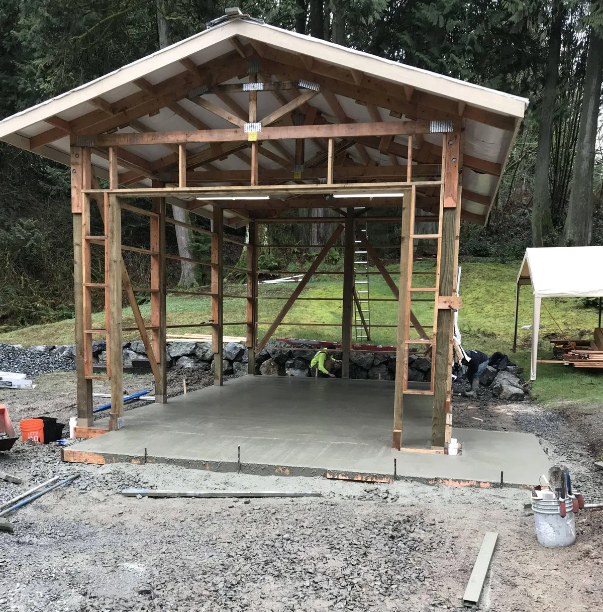Wood-framed pole building under construction over a fresh concrete slab with crushed-rock site preparation and drainage lines